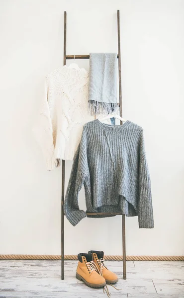 Fall or winter warm knitwear on hanger, white wall background. Fashionable female winter clothing. Woolen sweaters, grey scarf and winter boots
