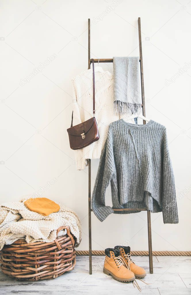 Fall or winter warm knitwear on hanger, white wall background. Fashionable female winter clothing. Woolen sweaters, scarf, boots, shoulder bag and knitted cap, copy space