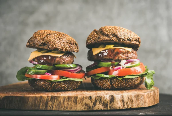 Beef meat cheeseburgers with barbeque sauce on rustic wooden board, grey concrete wall at background, close-up. Comfort food, burger bistro concept