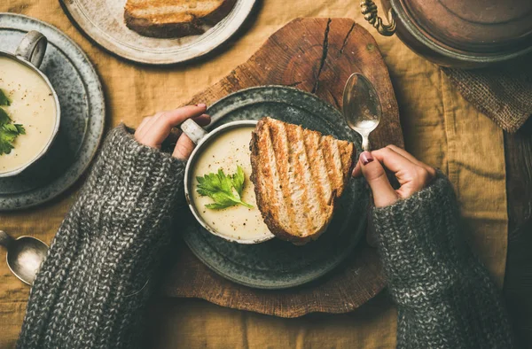 Autumn or Winter home dinner. Flat-lay of Fall warming celery cream soup and female hands with spoon over yellow linen tablecloth, top view. Comfort food, vegan, healthy and slow food concept