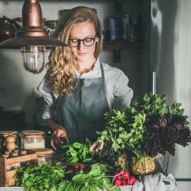 Fall seasonal vegetarian, vegan dinner cooking. Woman in apron and glasses cutting herbs and vegetables on concrete kitchen counter, square crop. Slow food, comfort food, healthy diet, clean eating
