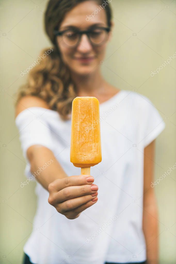 Healthy vegan orange mango citrus ice cream popsicle in hand of young woman with yellow wall at background, summer dessert and cheerful summer mood concept