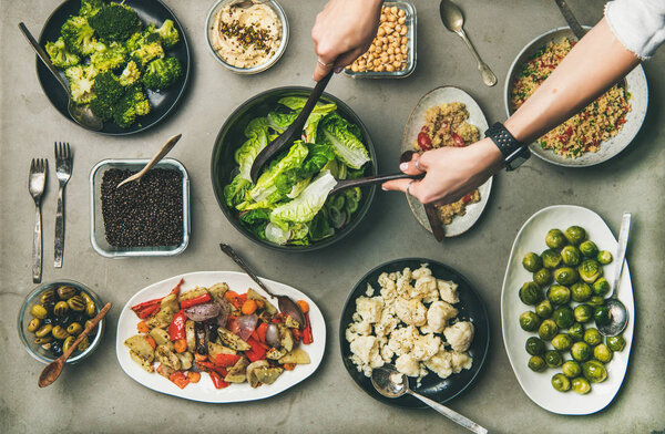Vegan dinner table setting. Healthy vegetarian dishes in plates on table. Flat-lay of vegetables, legumes, beans, olives, sprouts, hummus, couscous and female hands taking salad from bowl, top view