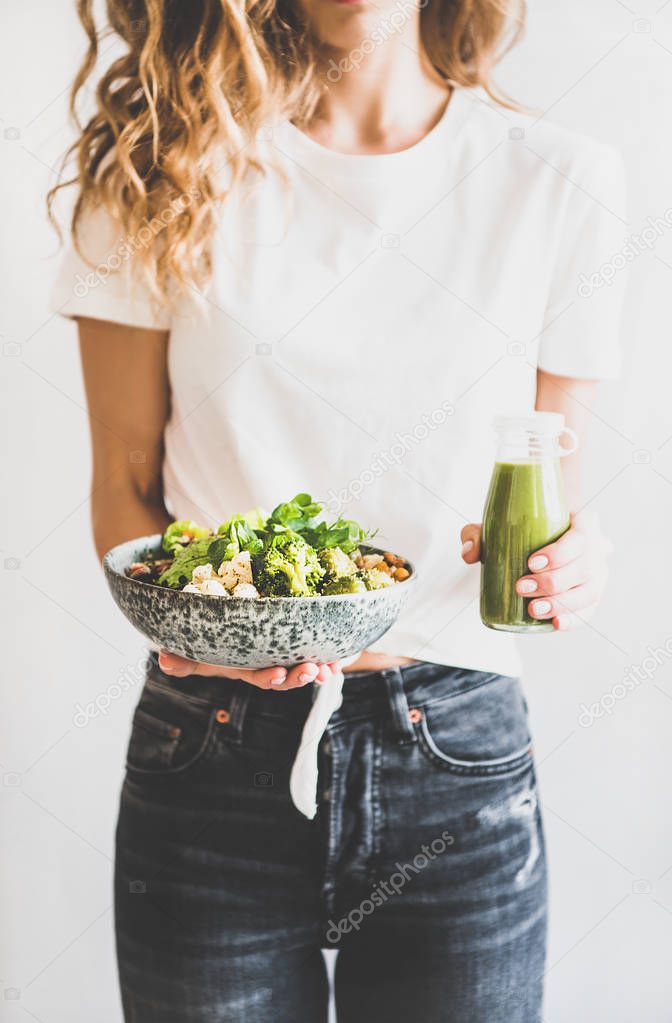 Healthy dinner, lunch. Woman in jeans standing and holding vegan superbowl or Buddha bowl with hummus, vegetable, fresh salad, beans, couscous and avocado and green smoothie in bottle