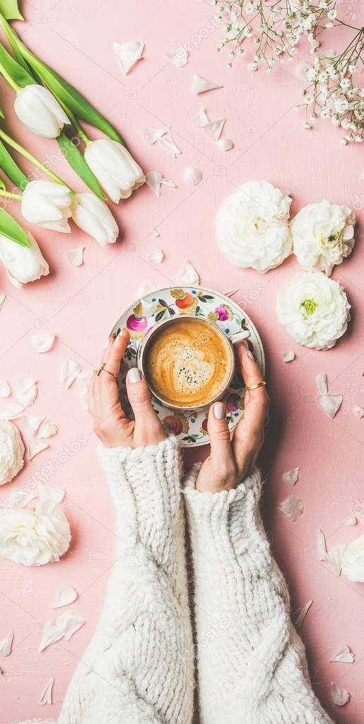 Flat-lay of female hands in knitted sweater holding cup of coffee, fresh white tulips and buttercup flowers over pink background, top view. Spring vibes or Womens Day holiday concept