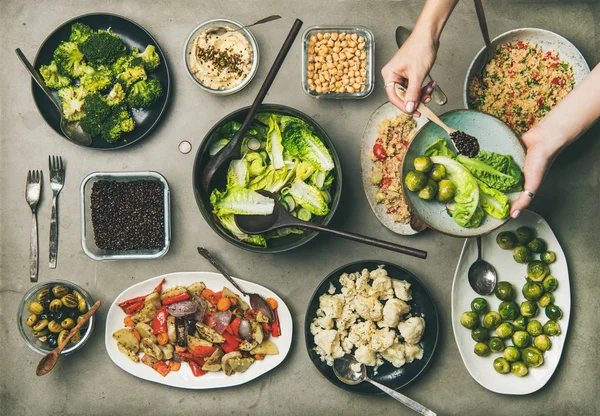 Vegan dinner table setting. Healthy dishes in plates on table. Flat-lay of vegetable salads, legumes, beans, olives, sprouts, hummus, woman hands adding ingredients to plate with spoon, top view