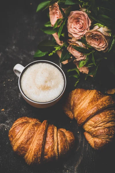 コーヒーブレイク 黒のみすぼらしい背景 トップビュー上の新鮮なクロワッサンやピンクの花の花束とホットカプチーノのカップのフラットレイ 春のムード — ストック写真