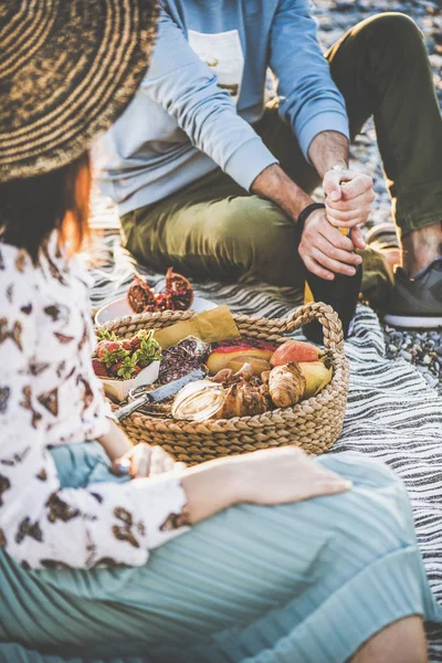 Summer beach picnic at sunset. Young couple having weekend picnic outdoors at seaside with fresh fruit and tray of tasty appetizers and opening bottle of sparkling wine