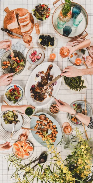 Family or friends gathering dinner. Flat-lay of hands of people eating lamb shoulder, salads, vegetables, drinking wine over checkered tablecloth, top view, vertical composition. Celebration party