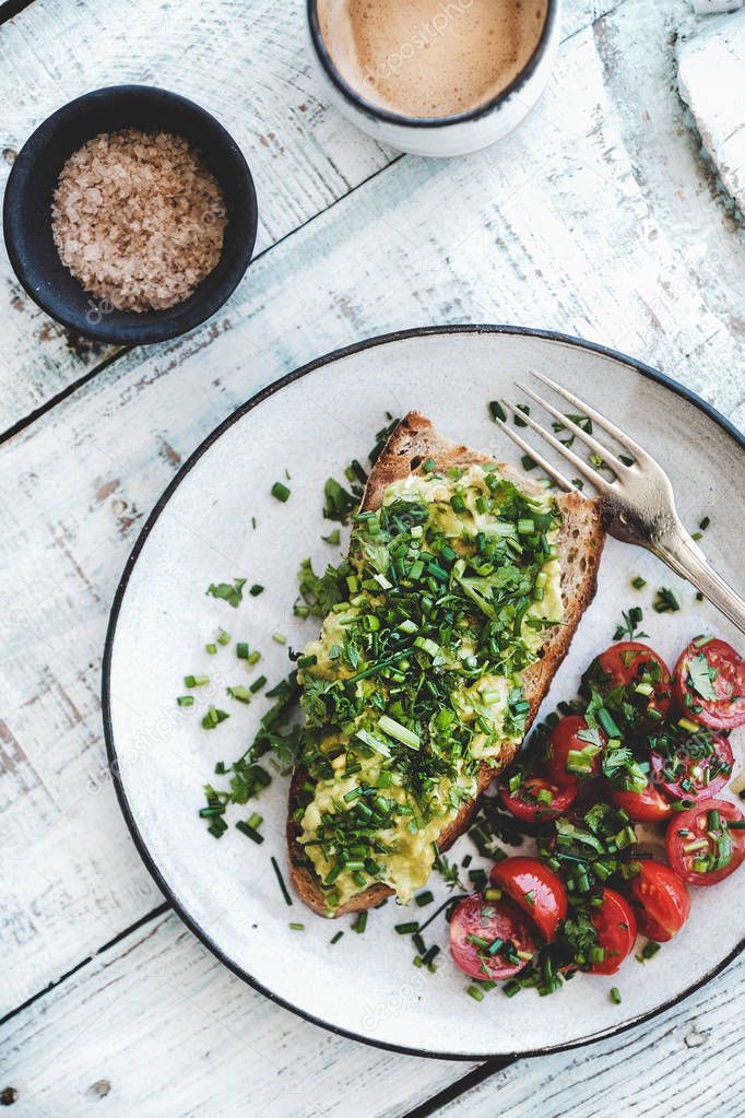 Healthy vegan breakfast. Flat-lay of avocado toast on sourdough bread with chives, coriander, cherry-tomatoes with cup of coffee over white background, top view. Vegetarian, clean eating concept