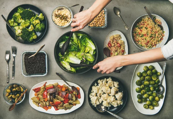 Vegan dinner table setting. Healthy vegetarian dishes in plates on table. Flat-lay of vegetables, legumes, beans, olives, sprouts, hummus, couscous and woman hands taking fresh salad from bowl, top view