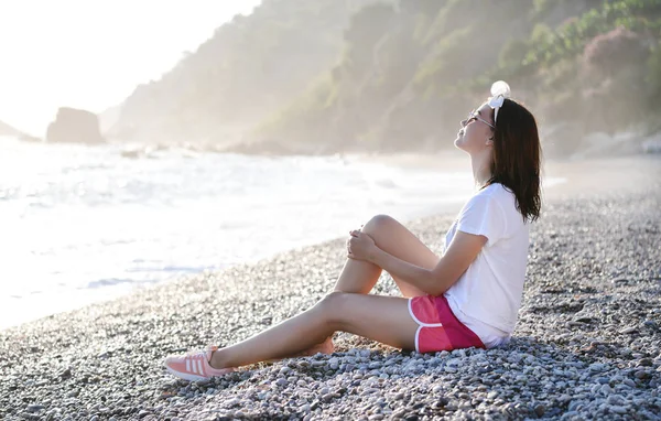 夏の美しい若い女性の外観と目を閉じて座って 地中海の小石の野生のビーチでリラックスしたサングラス スローリビングの概念 — ストック写真
