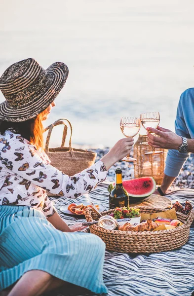 Summer beach picnic at sunset. Young couple having weekend picnic outdoors at seaside with fresh fruit and tray of tasty appetizers, drinking sparkling wine and enjoying lovely chat