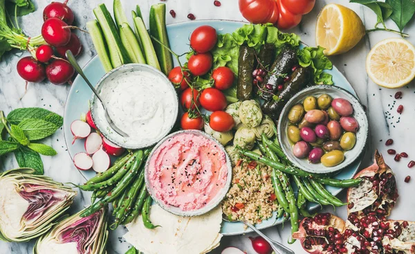 Healthy vegan snack set. Flat-lay of beetroot hummus, yogurt and herb dip, olives, flatbread, cous cous, dolma, vegetables and fruit on tray over marble background, top view. Clean eating food concept