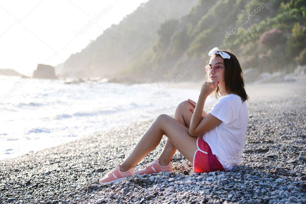 Beautiful young woman in summer look and sunglasses is dreaming and relaxing at pebble wild beach at Mediterranean sea. Traveling, tourism, vacation, freedom, slow living concept