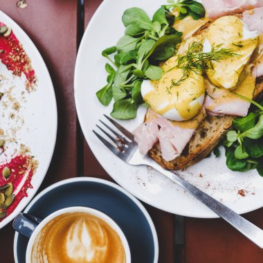 Trendy kahvaltı seti. Yeşil salata ve domuz jambonu ve kafe masada kahve fincanı ile ekşi hamur ekmek tost üzerinde haşlanmış Yumurta Benedict Düz yatıyordu, üst görünümü, kare ürün