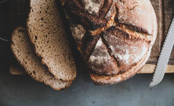 Flat-lay of freshly baked sourdough bread loaf and bread slices on wooden board over grey concrete table background, top view, copy space, close-up