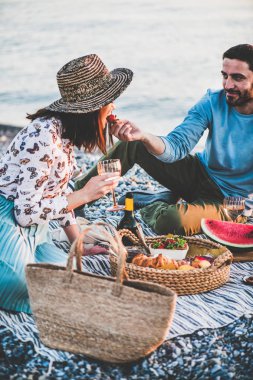 Gün batımında yaz plaj pikniği. Taze meyve ve lezzetli meze tepsiile deniz kenarında hafta sonu piknik açık havada olan genç çift, köpüklü şarap içme ve çilek ile birbirlerini besleme