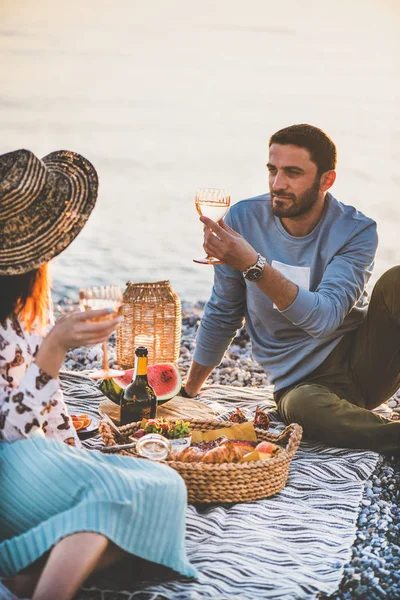 Summer beach picnic at sunset. Young happy couple having weekend picnic outdoors at seaside with sparkling wine, fresh fruit and tray of tasty appetizers, drinking wine and enjoying chat