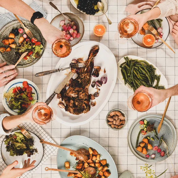 Family, friends gathering dinner. Flat-lay of peoples hands with glasses of rose wine over table with roasted lamb shoulder, salad, vegetables, mimosa branch, top view, square crop. Celebration party