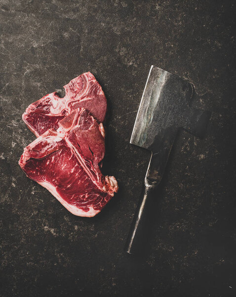 Flat-lay of fresh uncooked beef meat t-bone steaks and butchers cleaver over rough black stone background, top view. Butchers shop concept