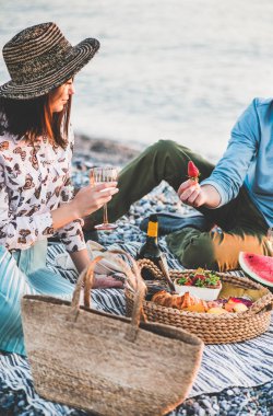 Gün batımında yaz pikniği. Genç çift hafta sonu deniz kenarında taze meyve ve lezzetli aperatiflerle piknik yapıyor, köpüklü şarap içiyor ve çilek yiyor.