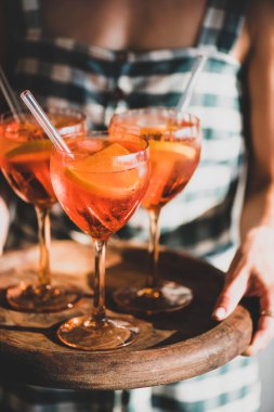 Kareli elbiseli genç bir kadın, elinde aperatif aperatif bardaklı Aperol Spritz tepsisiyle portakallı soğuk bir içecek, seçici bir odak noktası var. Yaz ferahlatıcı içecek konsepti