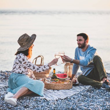 Gün batımında yaz pikniği. Genç mutlu çift hafta sonu pikniğini deniz kenarında şarap, meyve ve bir tepsi lezzetli aperatiflerle şarap içerek ve birbirine bakarak yapıyor.