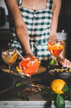 Damalı elbiseli genç bir kadın elinde Aperol Spritz aperatif kokteyli, mutfakta da portakallı ve camlı bir kokteyl tutuyor. Yaz ferahlatıcı içecek konsepti