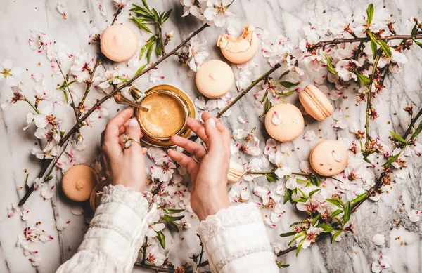 Flat-lay of womans hands over cup of hot espresso coffee, sweet macaron cookies, white spring blossom flowers and white marble background, top view