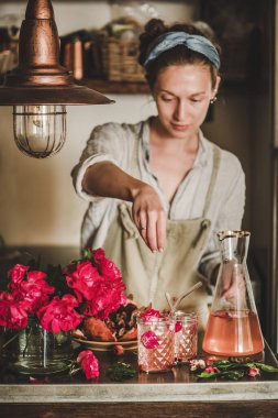 Keten önlüklü genç bir kadın, gül limonatasına buzlu ve beton mutfak tezgahının üzerinde taze pembe gül çiçekleri olan bardaklar ekliyor. Yaz ferahlatıcı soğuk içecek konsepti