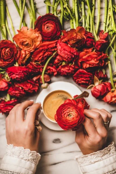 Flat-lay of womans hand holding red ranunculus flower over cup of black espresso coffee and white marble table woth flowers, top view. Seasonal spring or summer coffee break concept