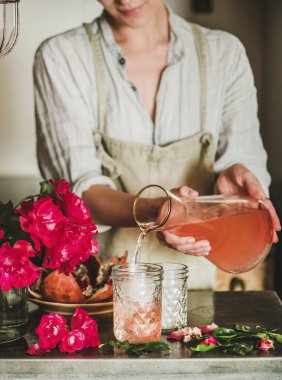 Keten önlüklü genç bir kadın taze pembe çiçeklerle sürahiden bardaklara kadar buzla gül limonatası dolduruyordu. Yaz ferahlatıcı soğuk içecek konsepti
