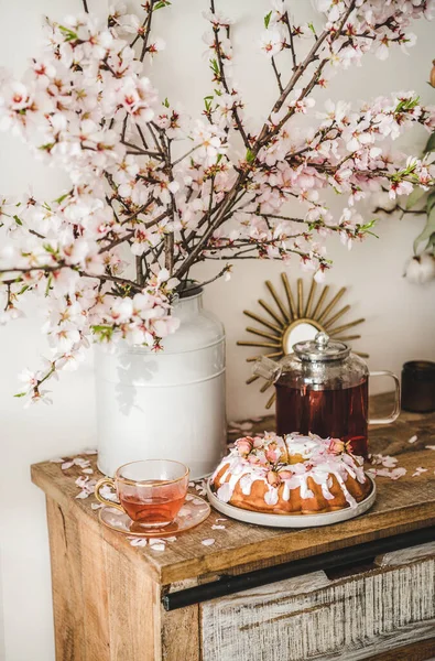 Spring or summer tea and cake setting. Rose and almond gluten-free bundt cake with rose flowers and black tea on rustic wooden cupboard under blooming branches in vase, white wall background