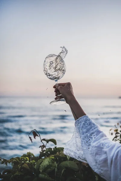 妇女们手握白色花边礼服袖子 玻璃杯白葡萄酒飘洒 背景是美丽的日落色和海平线 暑期野餐及小食饮品概念 — 图库照片