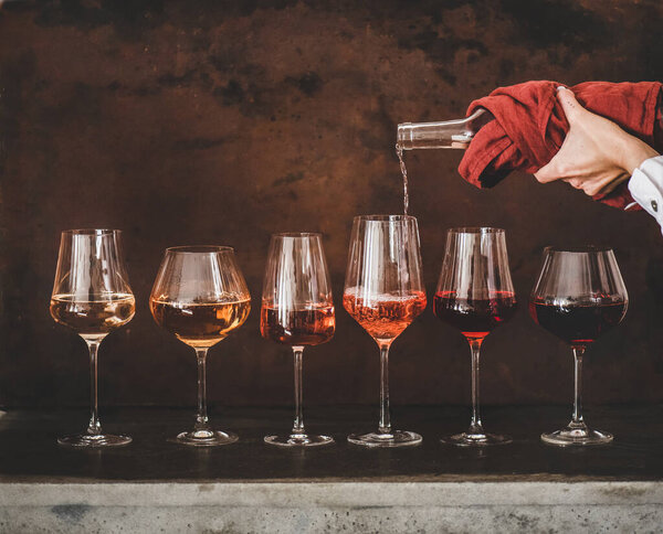 Shades of Rose wine in stemmed glasses placed in line and womans hand pouring wine from bottle to glass, rusty brown background behind. Wine bar, wine shop, tasting concept