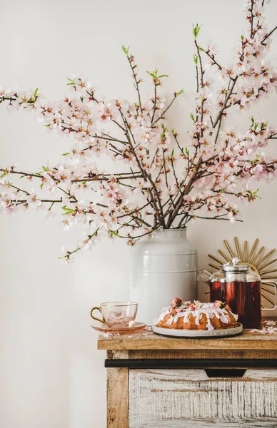 Spring or summer tea and cake setting. Rose and almond gluten free bundt cake with rose flowers and hot black tea on rustic wooden cupboard under blooming branches in vase, white wall at background
