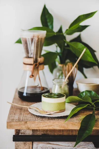 Coffee and dessert setup. Green matcha cheesecake and brewed pour-over coffee in glass flask on wooden kitchen counter, white wall background. Healthy, vegan, vegetarian, low calorie food concept