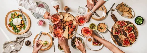 Friends wine and snacks party. Flay-lay of people hands clinking glasses with rose wine over table with cheese, fruit, smoked meat, tomato brushettas, buratta salad, top view. Wine tasting concept