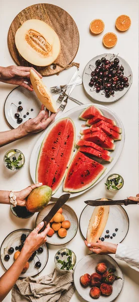 Summer tropical fruit party table. Flat-lay of lunch with berries, watermelon, lemonade and people cutting and holding fruits over white background, top view. Clean eating, fruiterian food concept