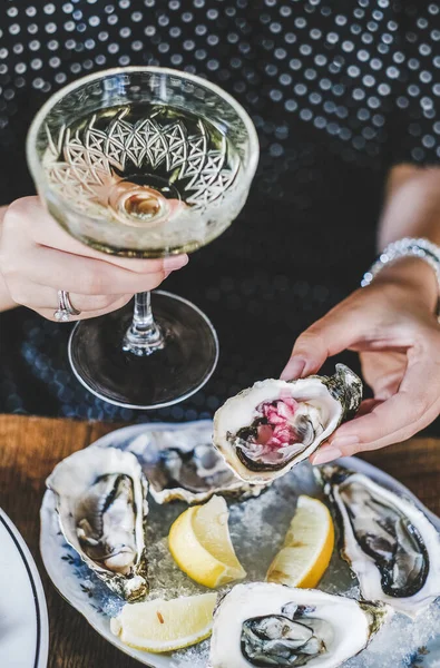 Hands of woman holding glass of champagne and fresh oyster with lemon juice in fish restaurant, selective focus. Seafood, French cuisine, fine dining concept
