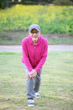Pembe parka giyen yaşlı Japon adam dışarıda bacak esnetiyor.