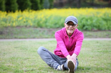 Kapüşonlu kıdemli Japon bir adam çimenlikte tek bacaklı öne doğru eğiliyor.