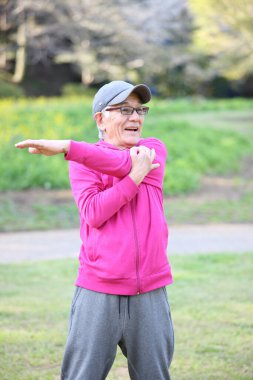  Pembe giysili yaşlı Japon adam dışarıda kol esnetiyor.
