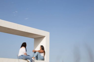 Genç arkadaşlar açık havada, açık havada mavi gökyüzü olan denize bakan bir parkın geometrik inşaatında oturarak konuşarak eğleniyorlar.