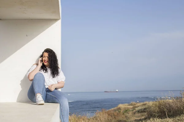 happy young woman laughs while talking on the phone sitting on a bench overlooking the sea