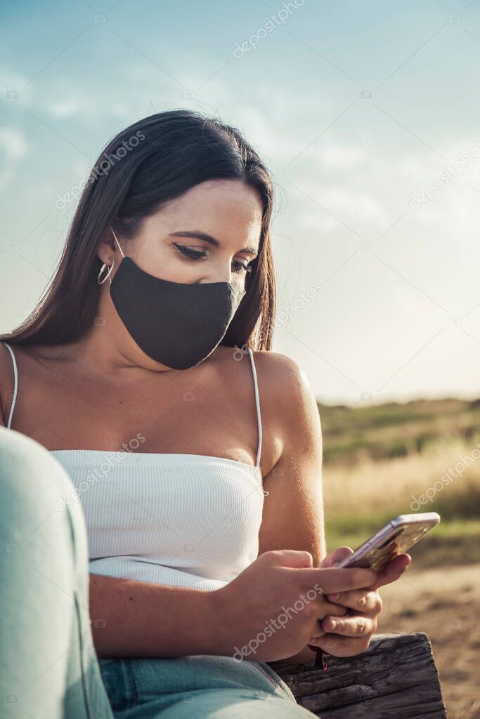 young woman with mask looks at her phone sitting on a bench in a park