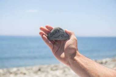 Bir el, üzerinde sörf yazan bir taş tutar. Arka planda plajı görebilirsiniz.