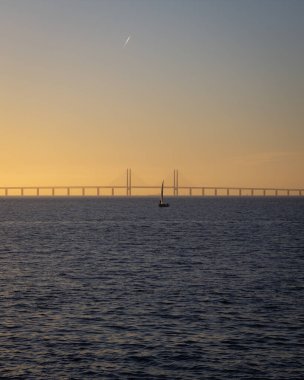 Günbatımında Danimarka ile İsveç arasında sakin bir okyanusta, arkasında Öresund Köprüsü olan tek bir yelkenli teknesi var..