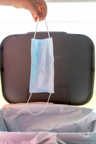 A hand depositing a surgical mask in the garbage can after use.